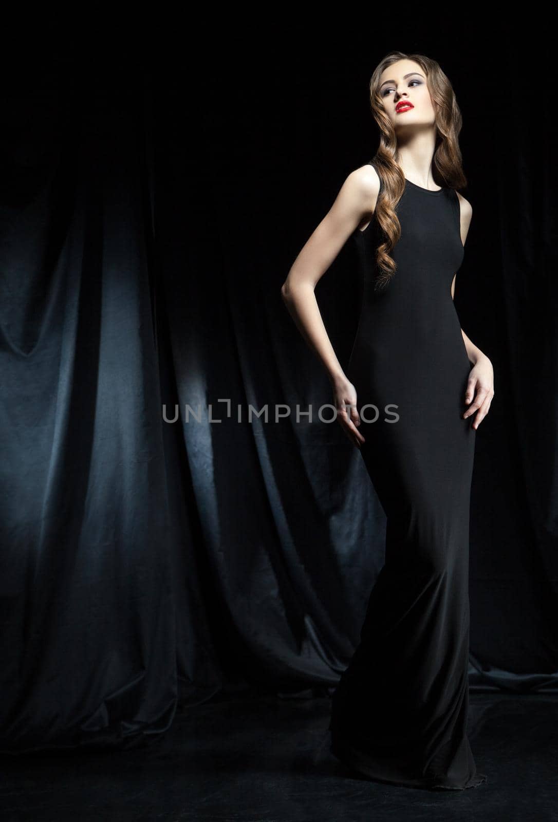 Portrait of young beautiful woman with long wavy hair in black dress posing against of black cloth