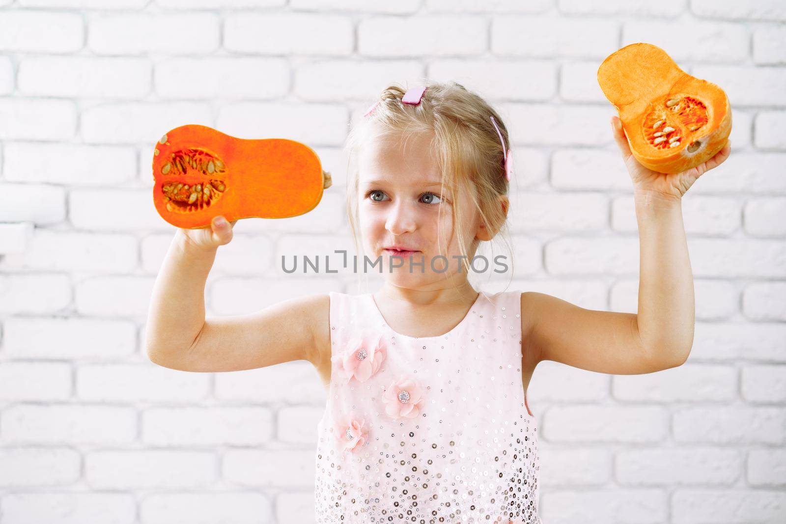 Cute little girl in pink dress holding pumpkin in her hands against white brick wall