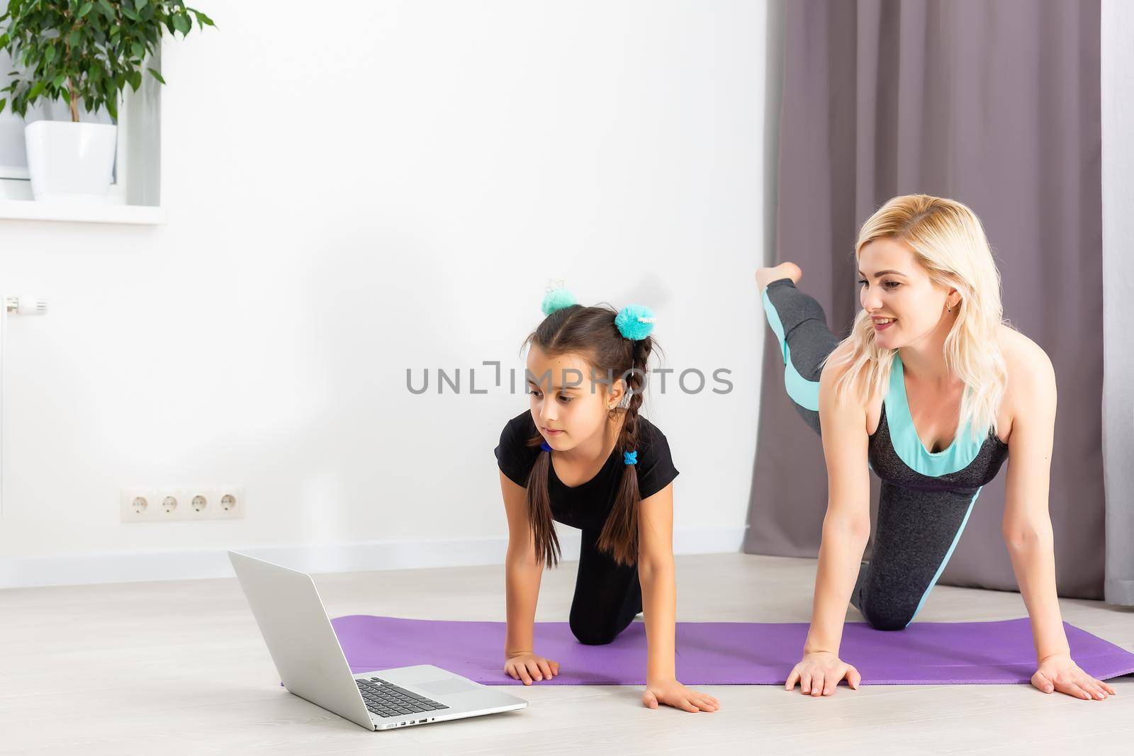 Mom and little daughter are doing gymnastics on the mat at home. They do yoga. They are fun because they have a happy family. Poses they are looking at the laptop. by Andelov13