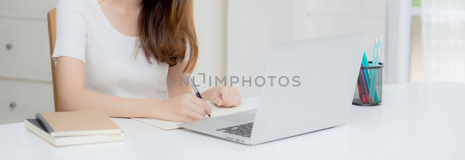 Young asian woman writing on notebook for planning working and using laptop computer on desk at home, girl notes about finance, female study and learning, business and communication concept.