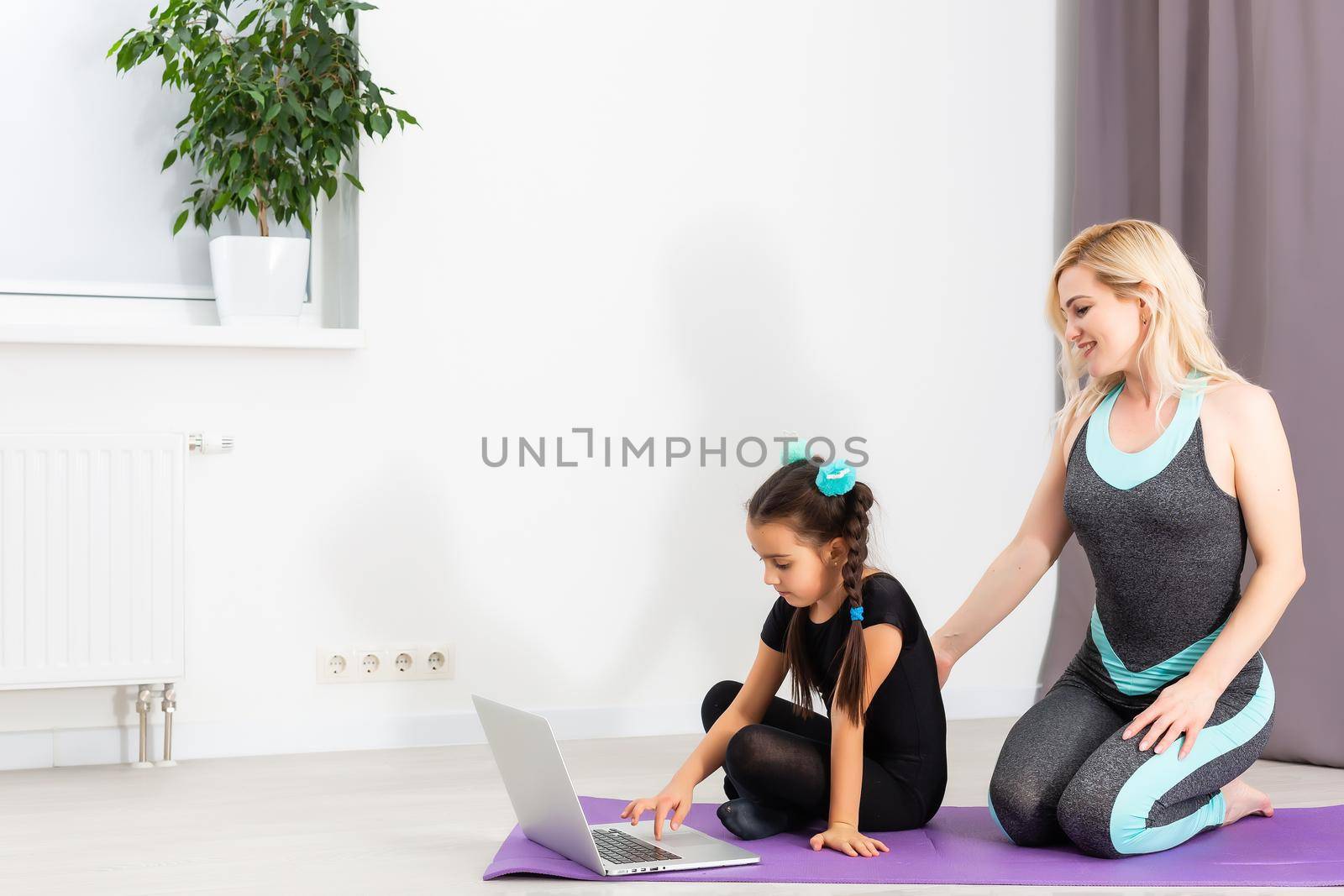 Mom and little daughter are doing gymnastics on the mat at home. They do yoga. They are fun because they have a happy family. Poses they are looking at the laptop. by Andelov13