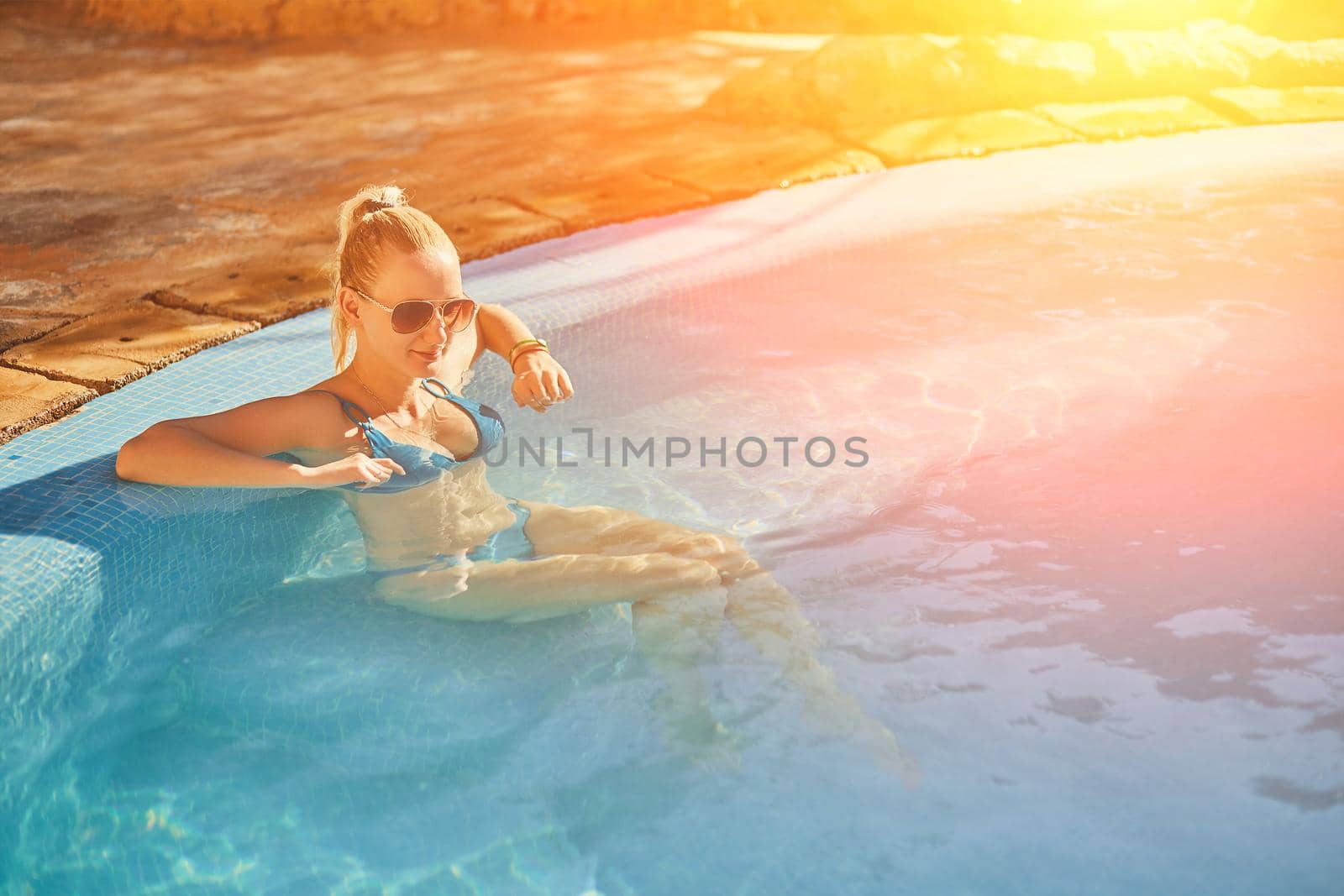 Woman in blue swimsuit and sunglasses relaxing in outdoor pool with clean transparent turquoise water. Woman sunbathing in bikini at tropical resort. Sun flare