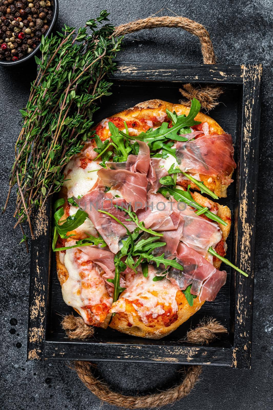 Italian Pizza with prosciutto parma ham, arugula salad and cheese in a rustic wooden tray. Black background. Top view by Composter