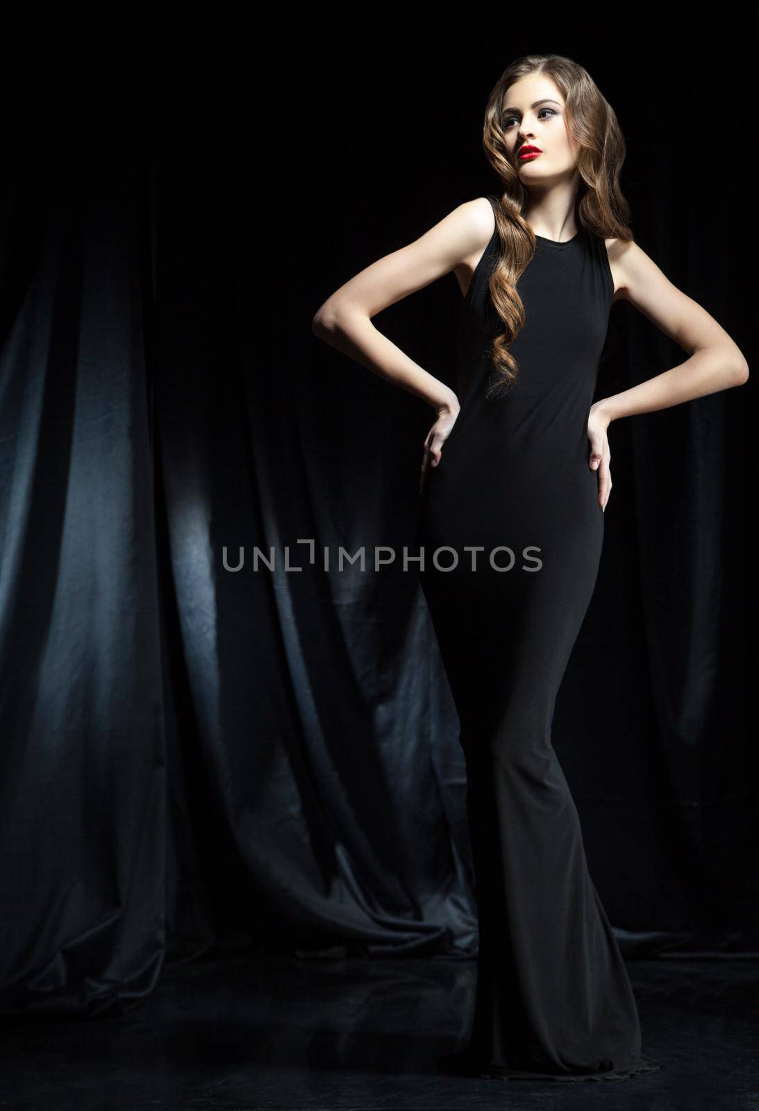 Portrait of young beautiful woman with long wavy hair in black dress posing against of black cloth
