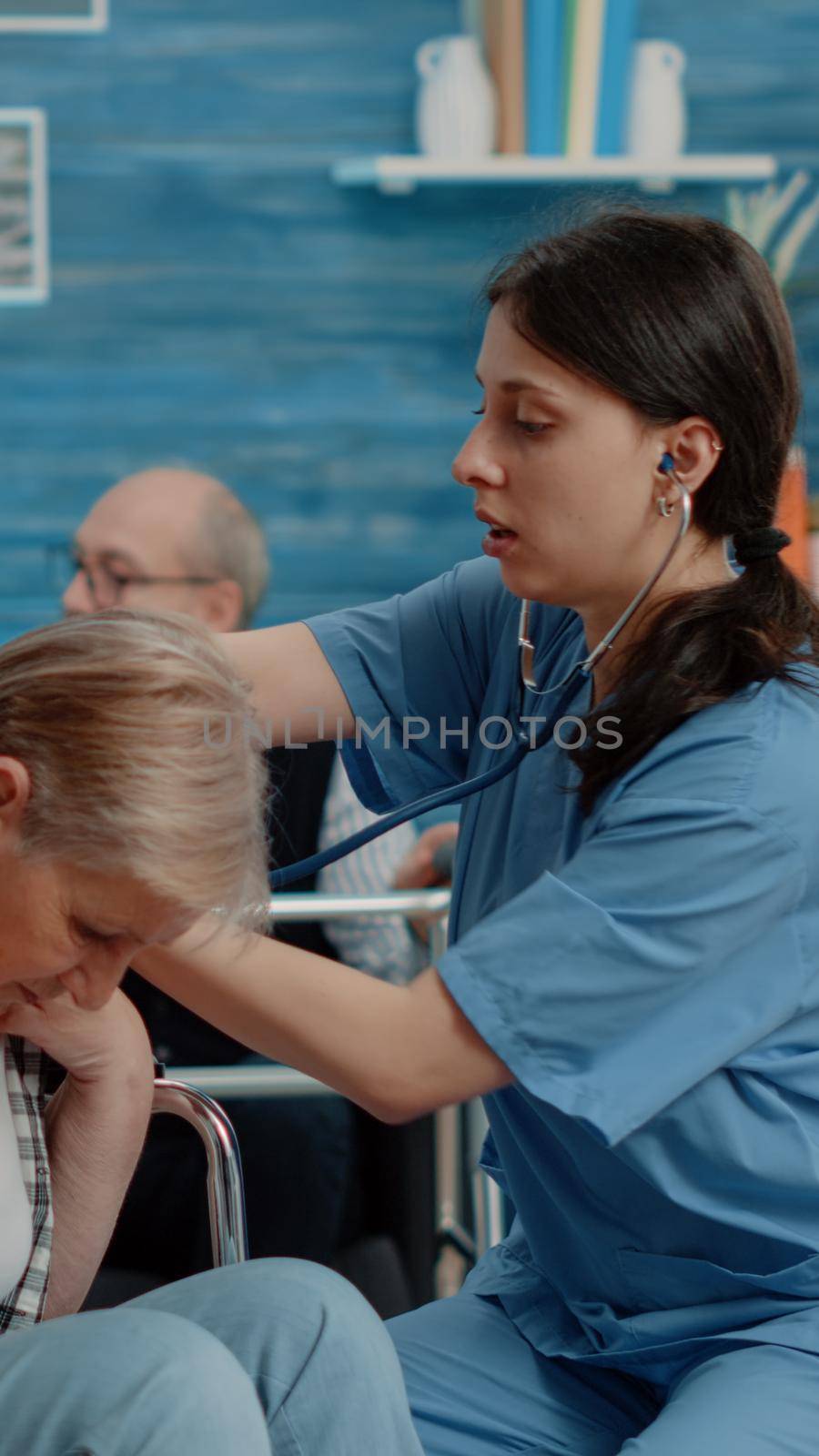 Medical assistant using stethoscope on senior patient in nursing home. Nurse checking pulse and heartbeat of retired woman with disability sitting in wheelchair for healthcare checkup.