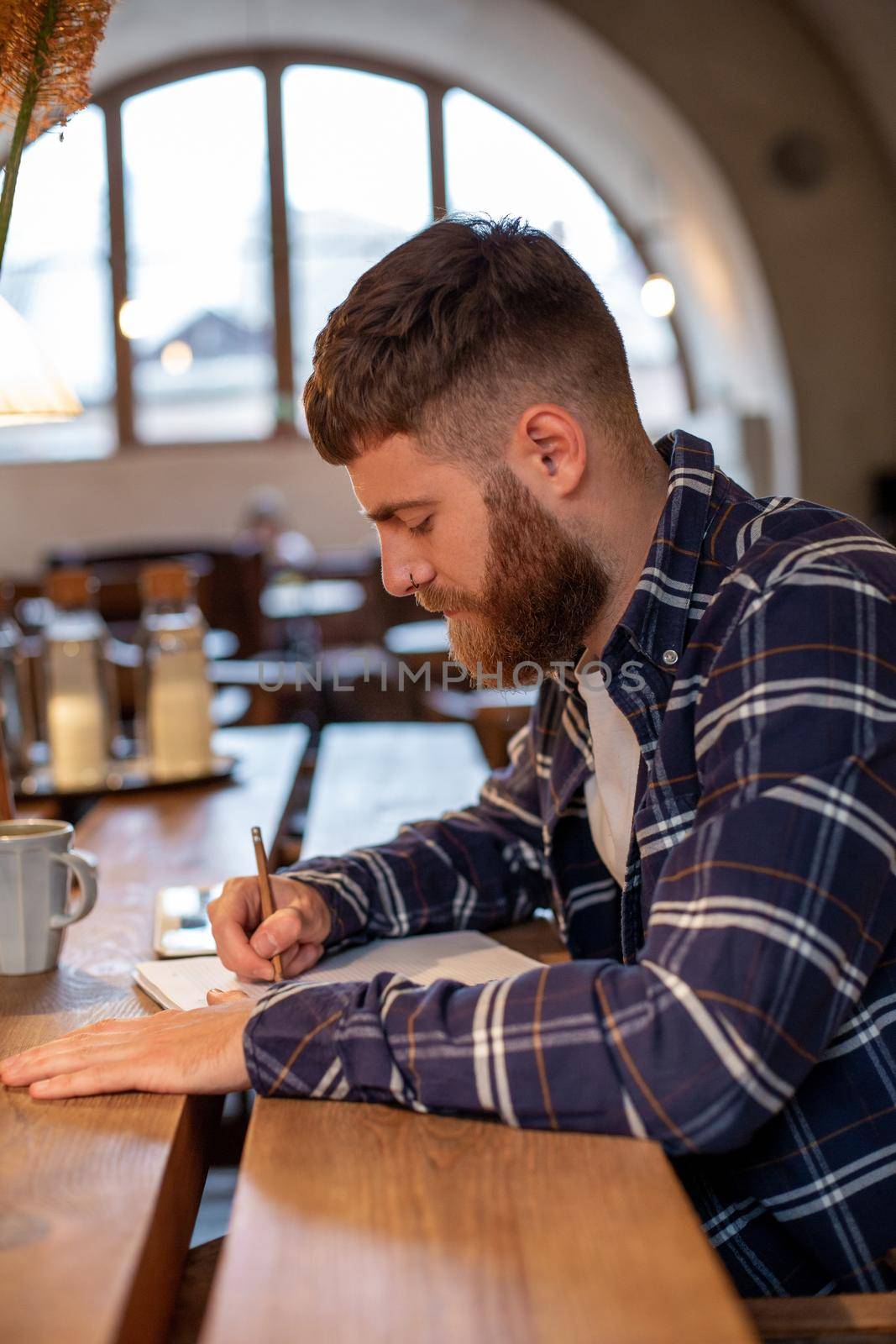 Young bearded businessman sits in cafe, home at table and writes in notebook, near lies tablet computer. Man is working, studying. by nazarovsergey