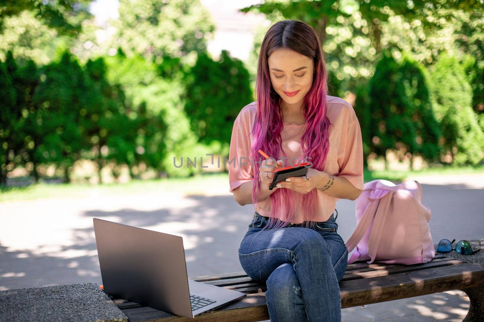 Woman writing in a notebook sitting on a wooden bench in the park. Girl working outdoors on portable computer, copy space. Technology, communication, freelance and remote working concept.