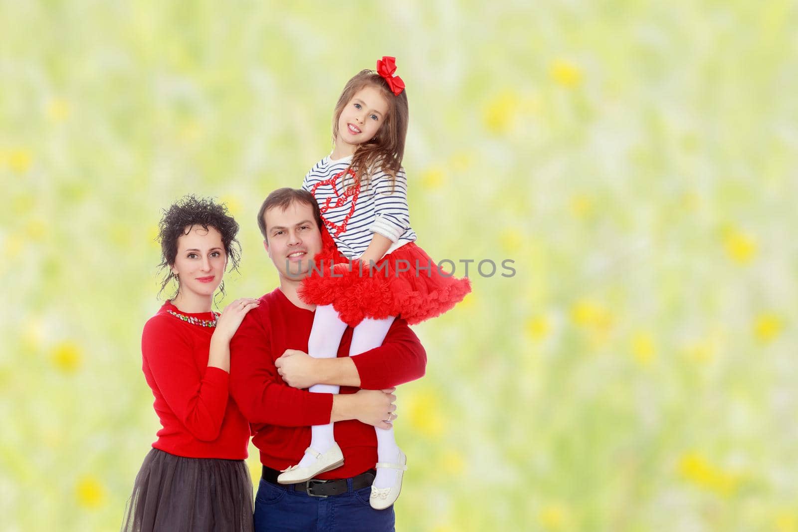 Happy young family dad mom and a little girl in bright red outfits . Dad holds daughter on hands.Summer white green blurred background.
