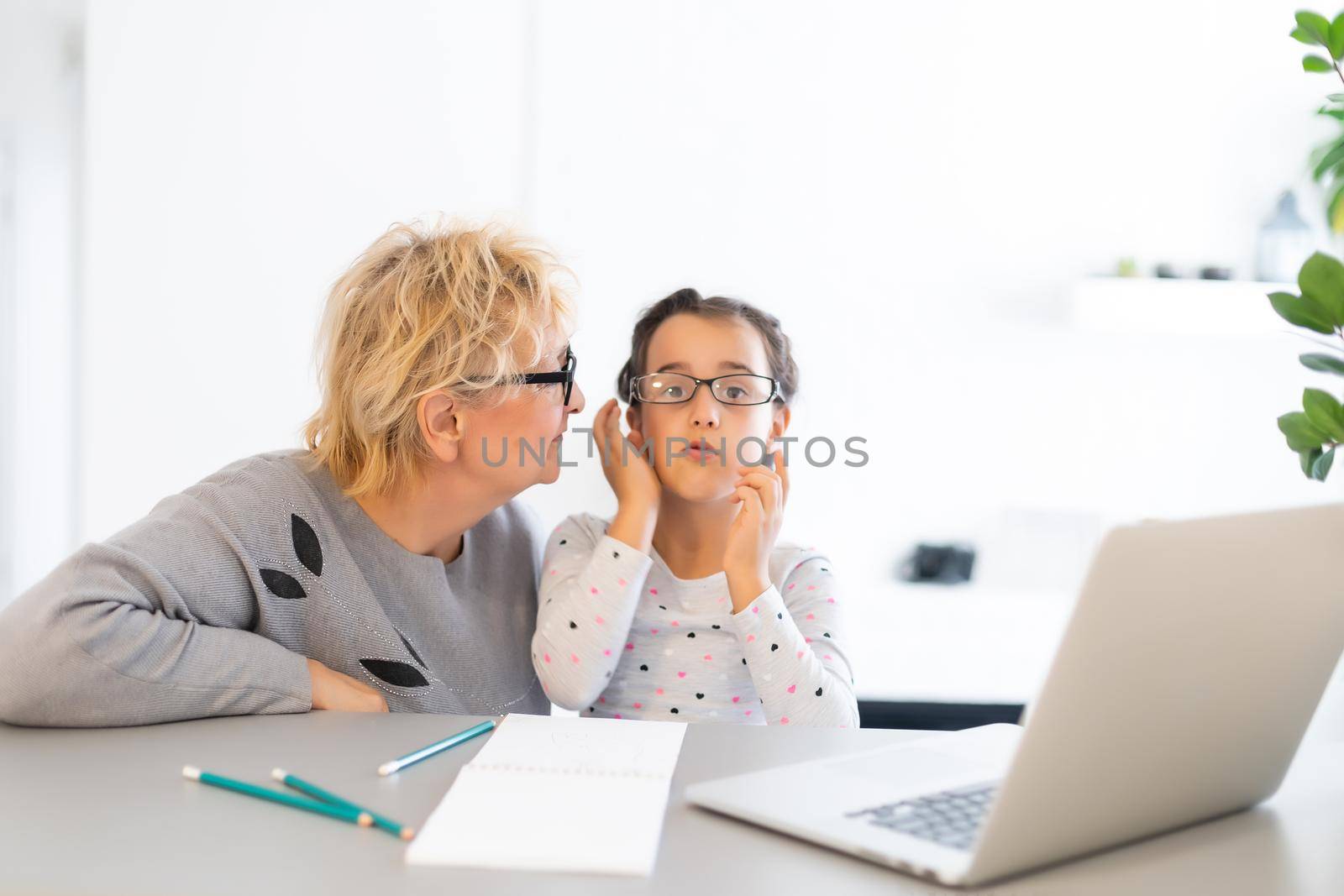 Mature grandmother helping child with homework at home. Satisfied old grandma helping her granddaughter studying in living room. Little girl writing on notebook with senior teacher sitting next to her