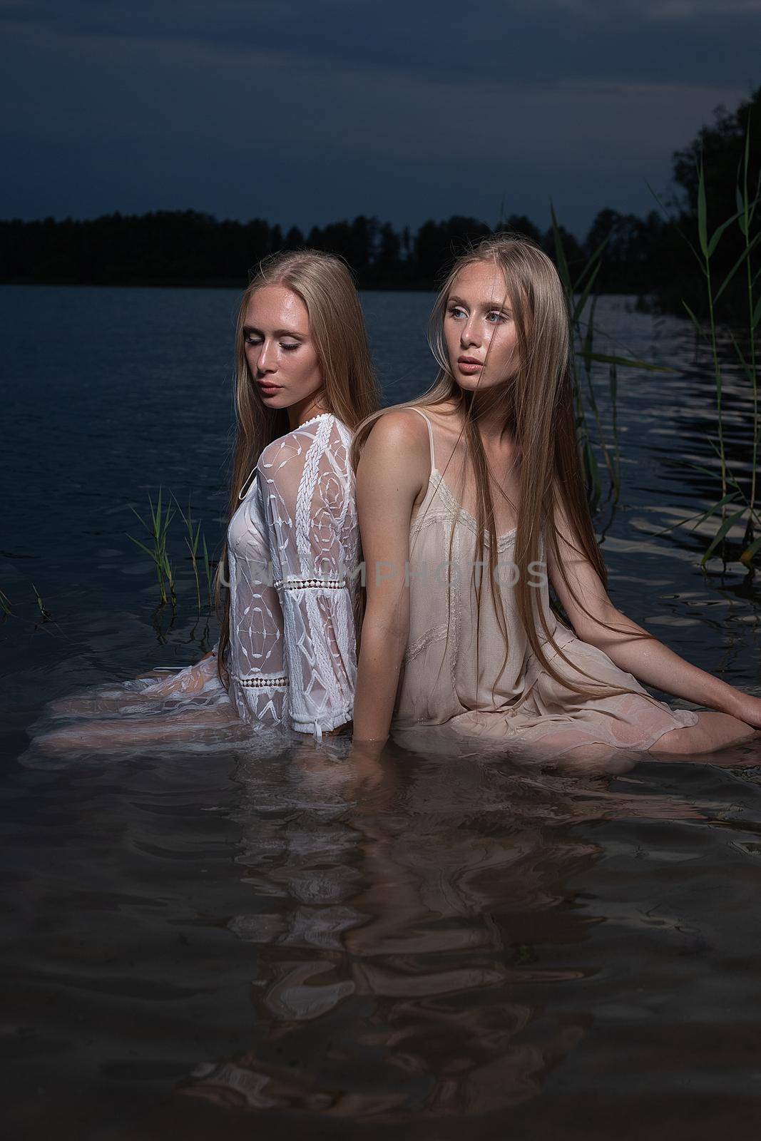 two attractive young twin sisters with long blond hair posing in light dresses in water of lake at summer night. stylish fashion photoshoot with flashlight. pretty models outdoors evening photosession