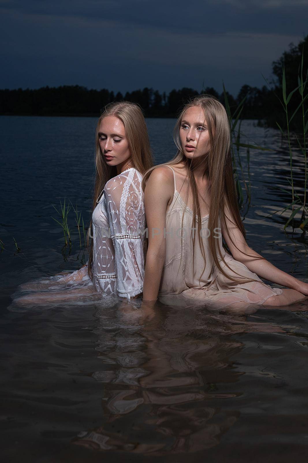 two young twin sisters posing in light dresses in water of lake at summer night by artemzatsepilin