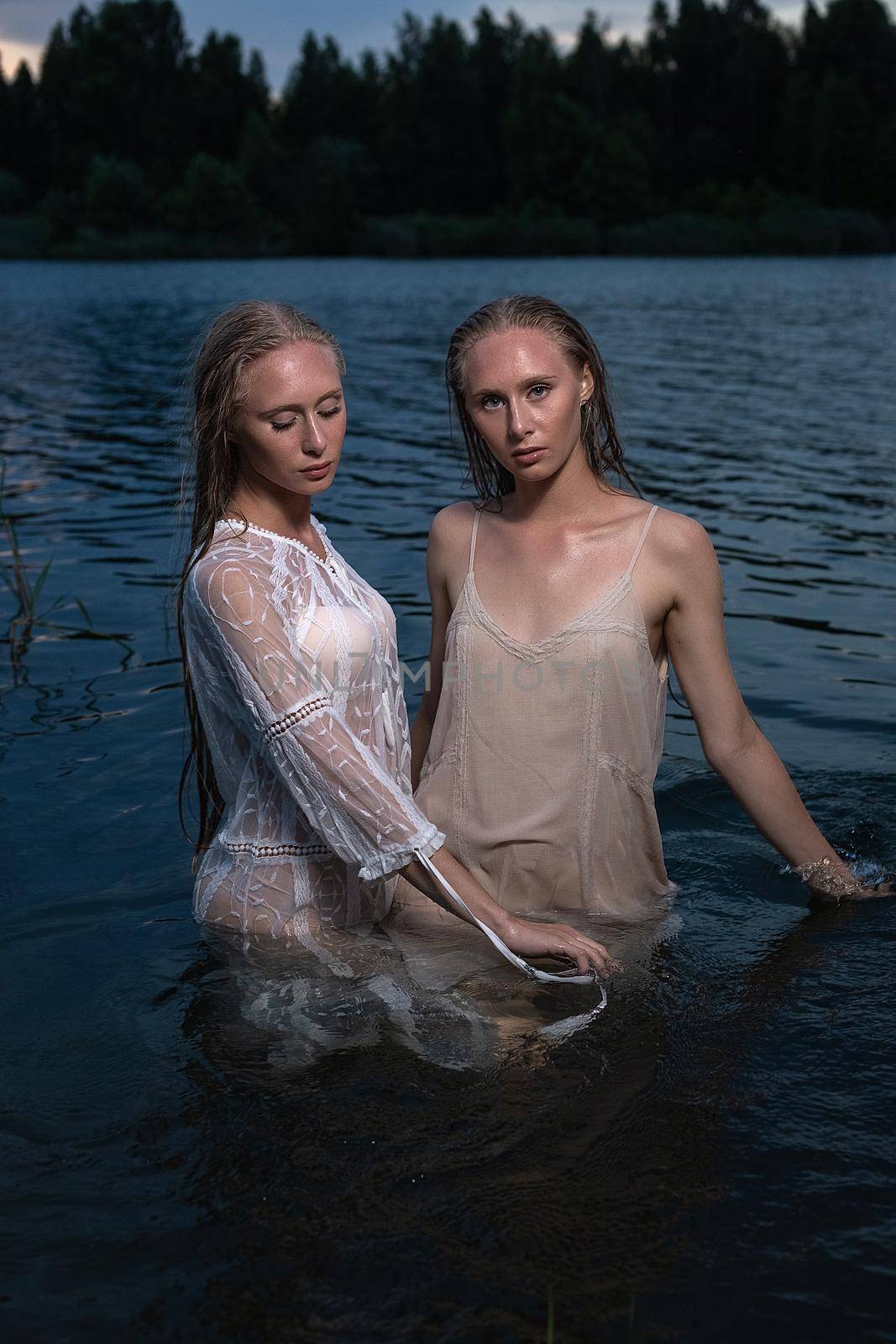 two young twin sisters with long blond hair posing in light dresses in water of lake at summer night, looking at camera. stylish fashion photoshoot with flashlight. outdoors evening photosession