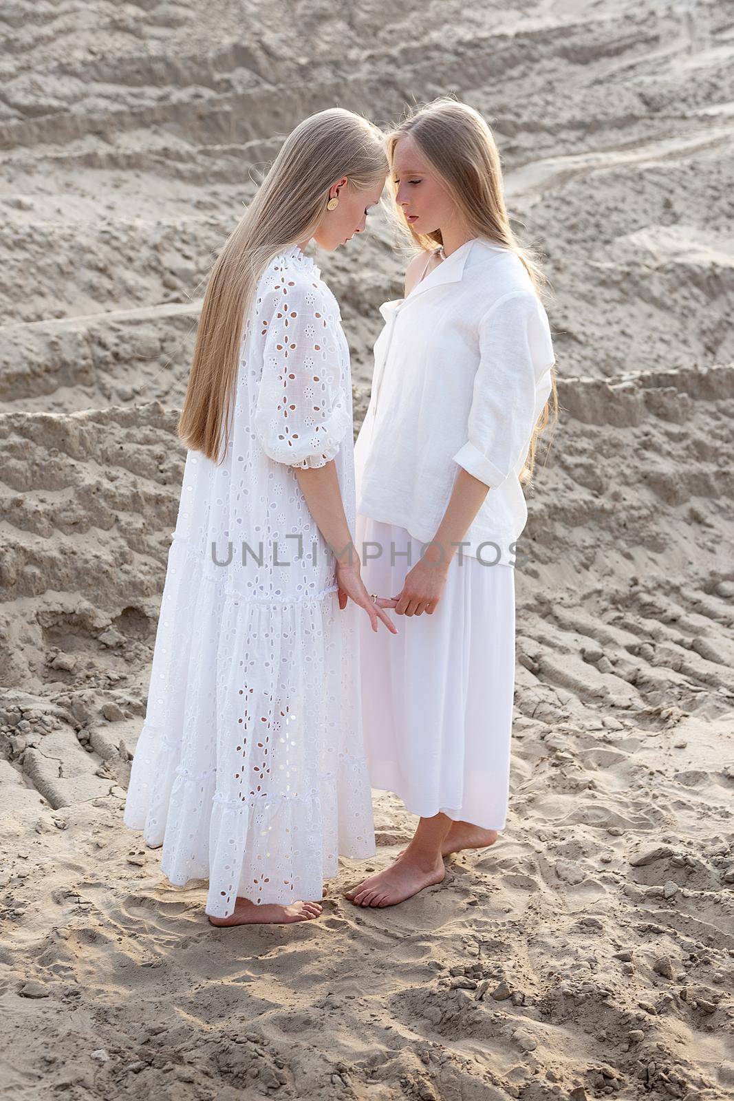 young pretty twins with long blond hair hold hands at sand quarry in elegant white dress, skirt, jacket. stylish fashion photoshoot, summer photosession. identical sisters spend time together outdoors