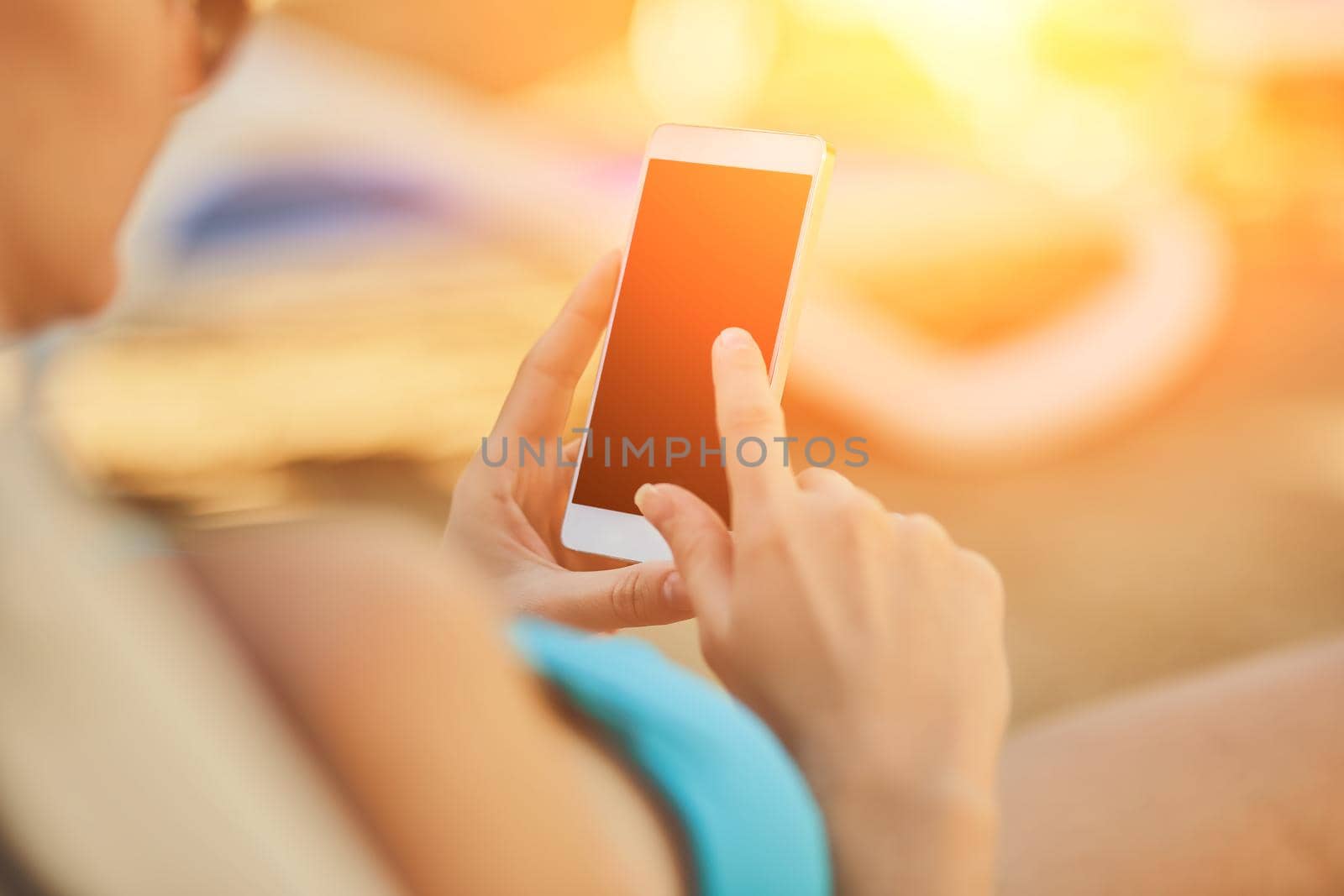 Close up view of a young attractive woman on holiday laying down on a white chaise-longue, holding and using a smartphone. Black display. People travel technology. Sun flare