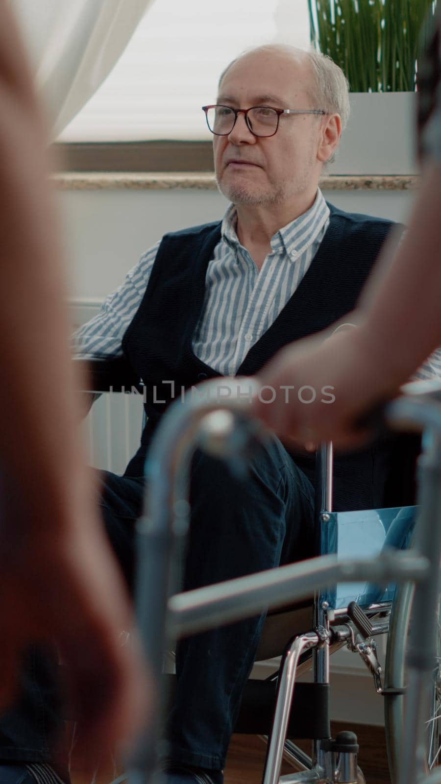 Old disabled man relaxing in nursting home room. Retired person with disability sitting in wheelchair preparing for recovery and wellness while nurses helping patient with walk frame