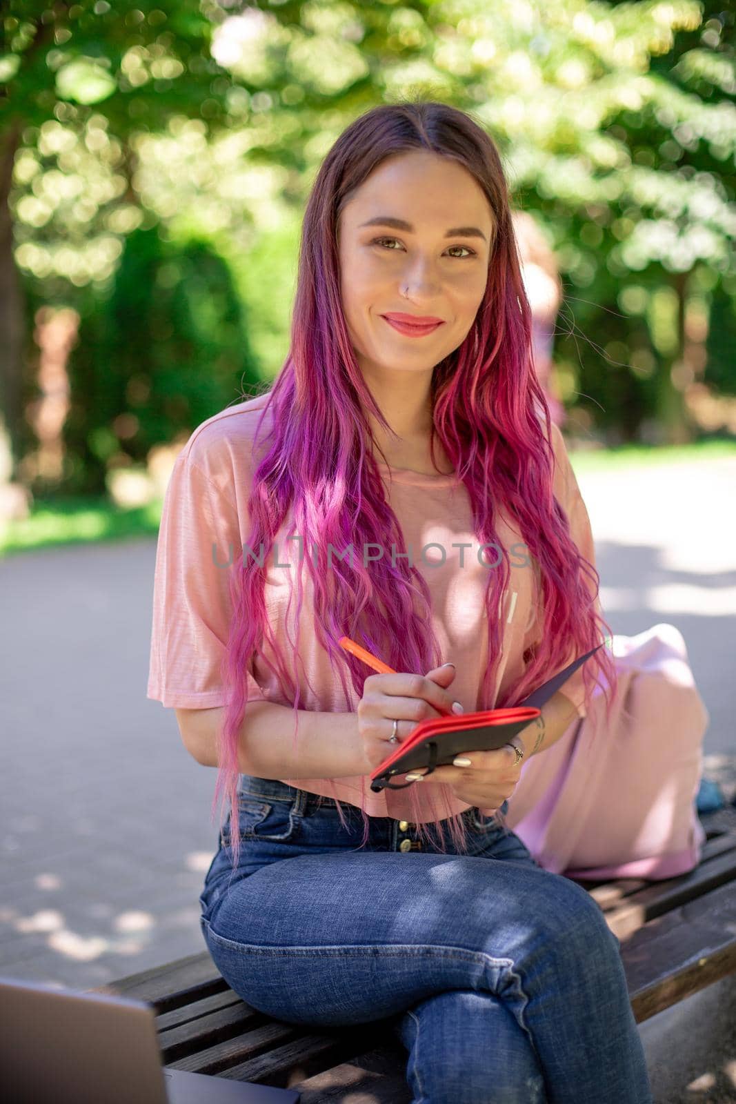 Woman writing in a notebook sitting on a wooden bench in the park. Girl working outdoors on portable computer, copy space. Technology, communication, freelance and remote working concept.