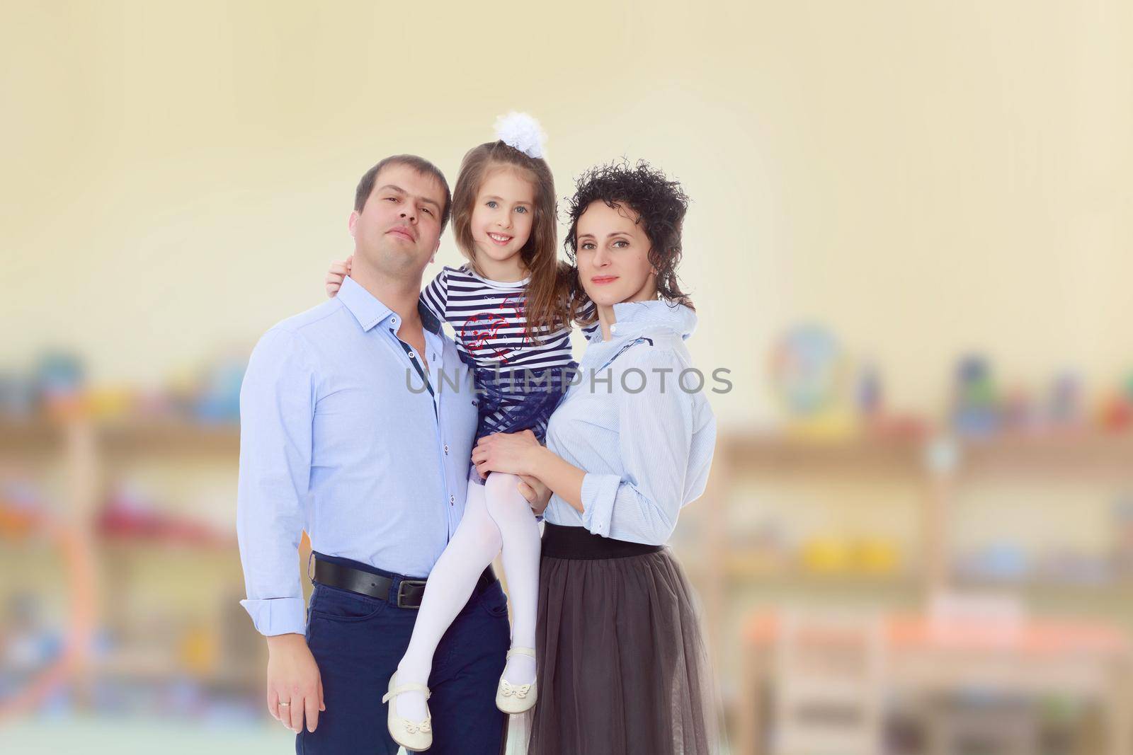 Happy young family, mom dad and little daughter.Parents keep the girl in her arms , and she hugs their neck.In the Montessori room the children's garden where there are shelves with toys and material.