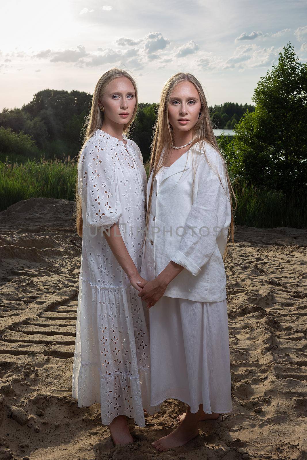 young twins with long blond hair posing at sand beach in elegant white clothes by artemzatsepilin