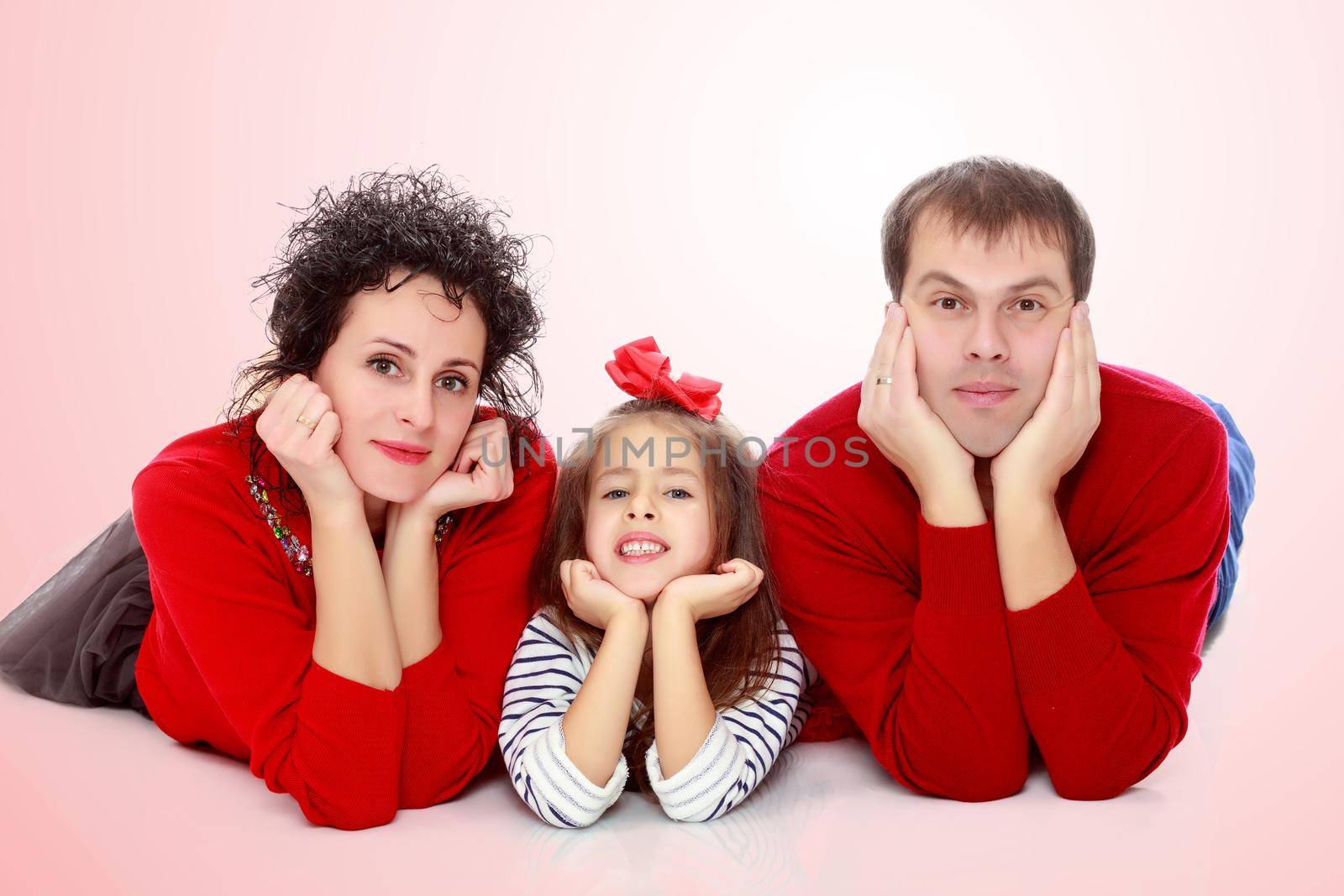 Happy young family dad mom and a little girl in bright red outfits . Family lying on the floor leaning on his hands.Pale pink gradient background.