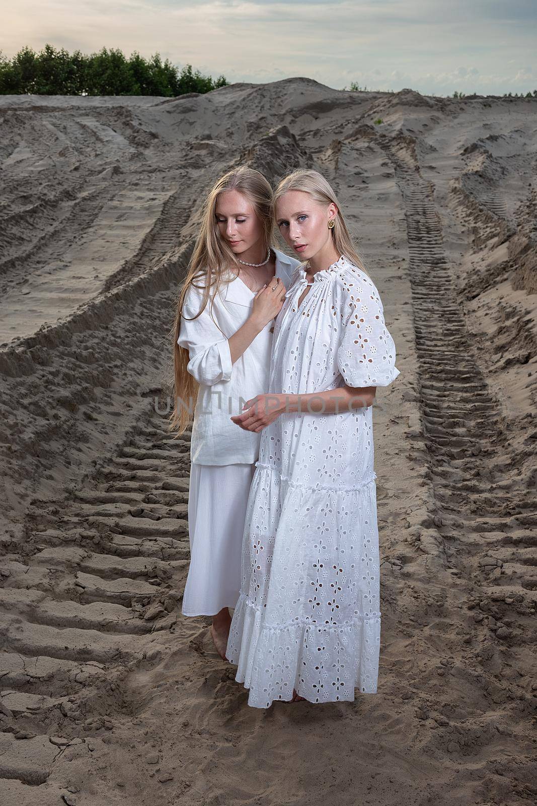 young blond pretty twins posing at sand quarry in elegant white dress, skirt, jacket. stylish fashion photoshoot with flashlight, summer photosession. identical sisters spend time together outdoors