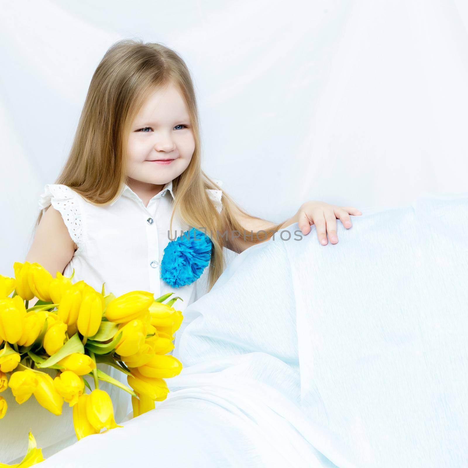 Cute little girl with long blond hair in a long blue skirt sitting on the couch with a large bouquet of bright yellow tulips.