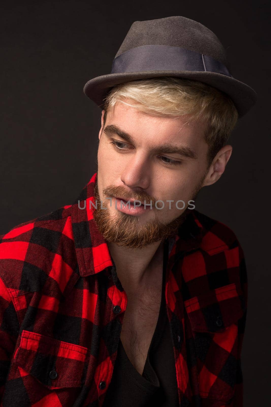 Close-up portrait of attractive young bearded hipster man wearing hat dressed in shirt in a cage isolated over black background. Studio shot