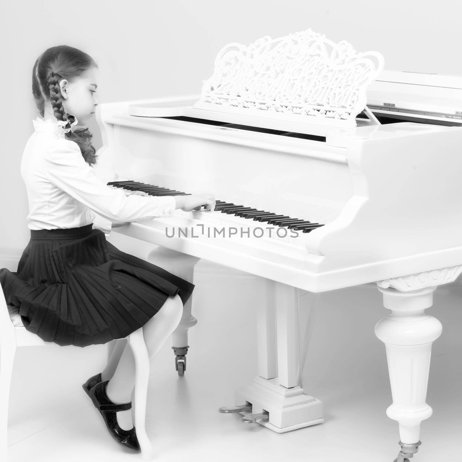 Charming little blonde with long wattled hair in plaits, playing on a white grand piano. In musical school.Black-and-white photo. Retro style.