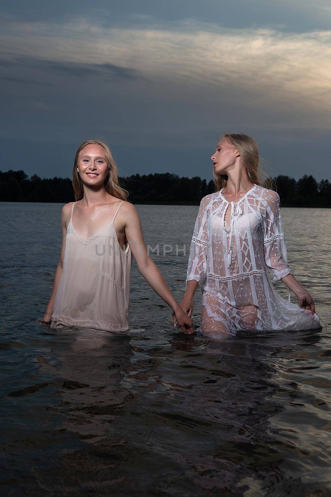 two young twin sisters posing in light dresses in water of lake at summer night by artemzatsepilin