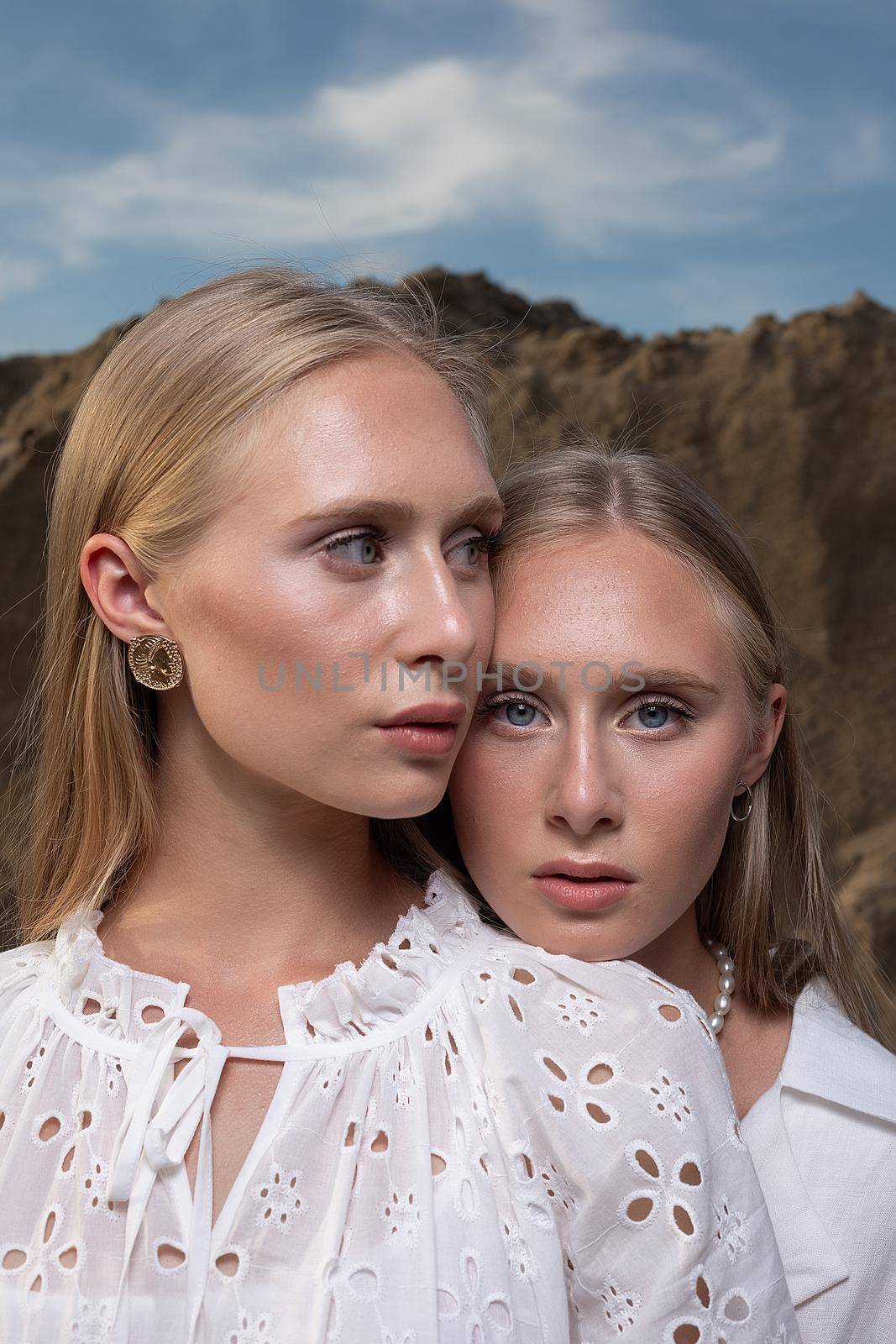 two young blond pretty twins posing at sand quarry in elegant white clothes by artemzatsepilin