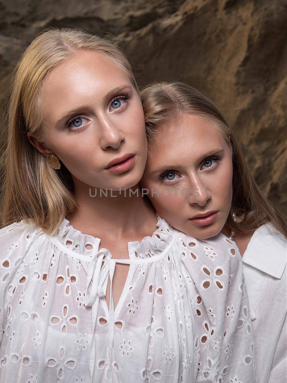 two young blond pretty twins posing at sand quarry in elegant white clothes. stylish glamorous fashion photoshoot with flashlight. girl holds her head on shoulder of her sister in front of sand wall