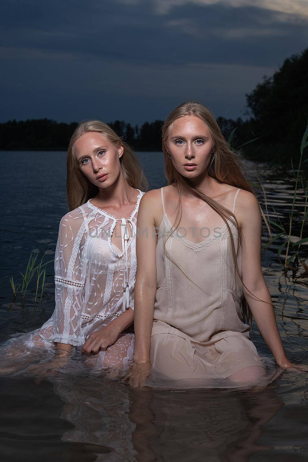 two young twin sisters with long blond hair posing in light dresses in water of lake at summer night, looking at camera. stylish fashion photoshoot with flashlight. outdoors evening photosession