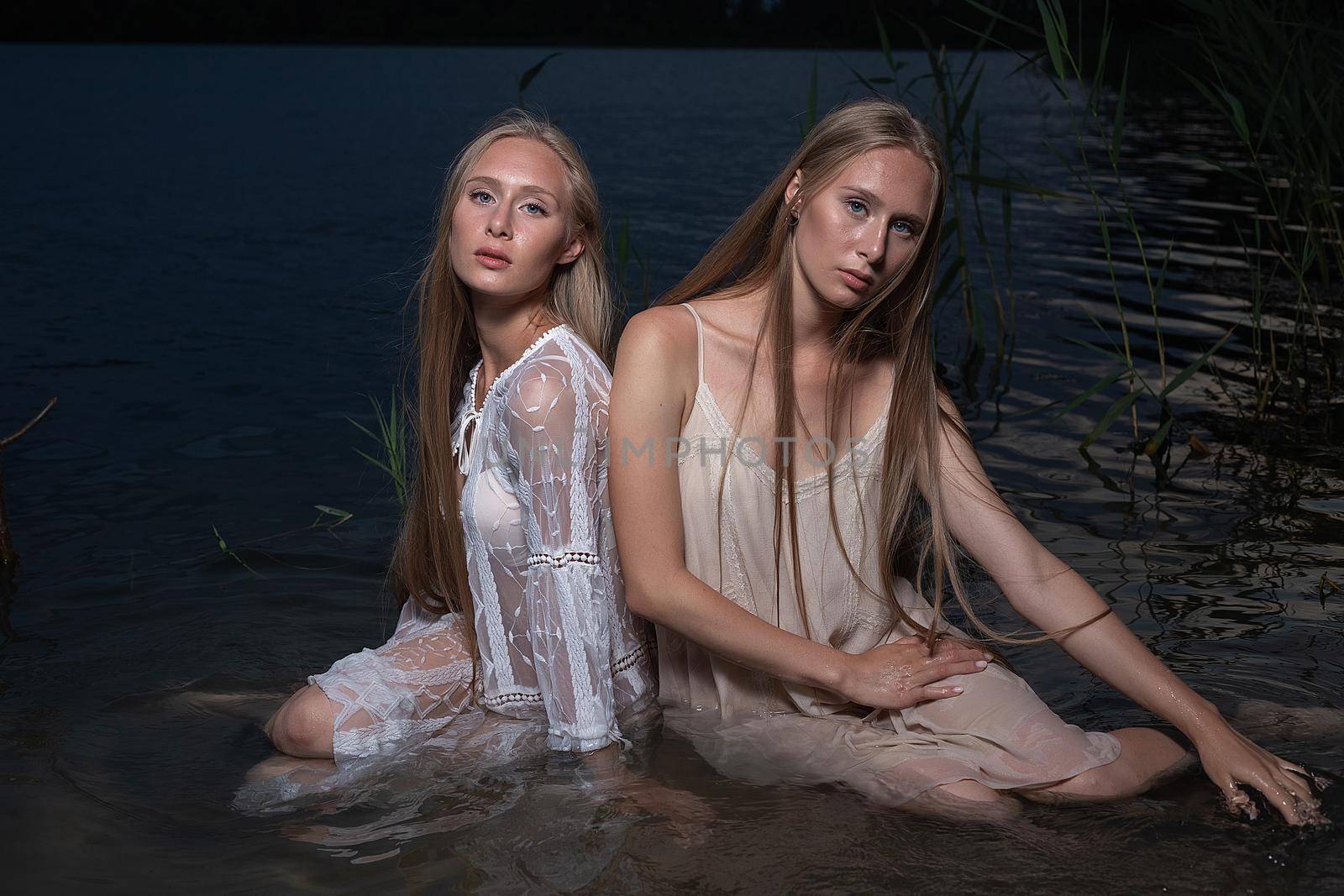 two young twin sisters with long blond hair posing in light dresses in water of lake at summer night, looking at camera. stylish fashion photoshoot with flashlight. outdoors evening photosession