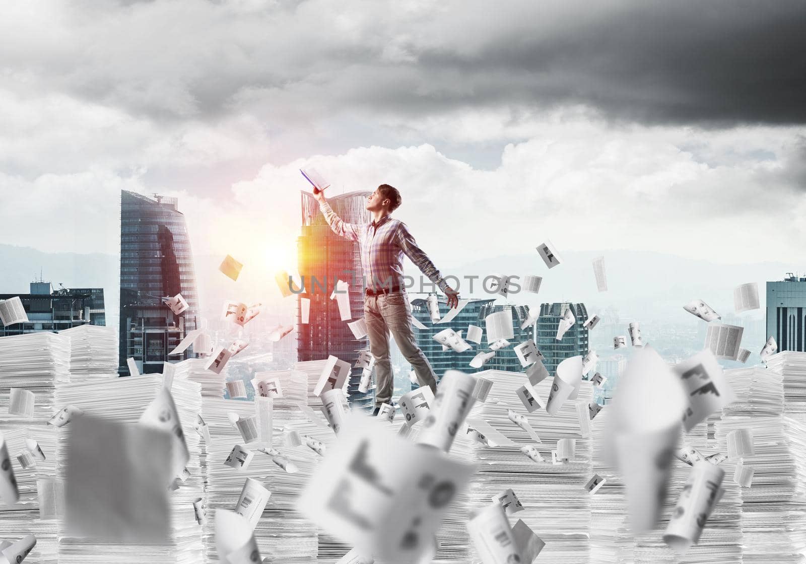 Side view of man in casual wear keeping hand with book up while standing among flying papers with cityscape and sunlight on background. Mixed media.