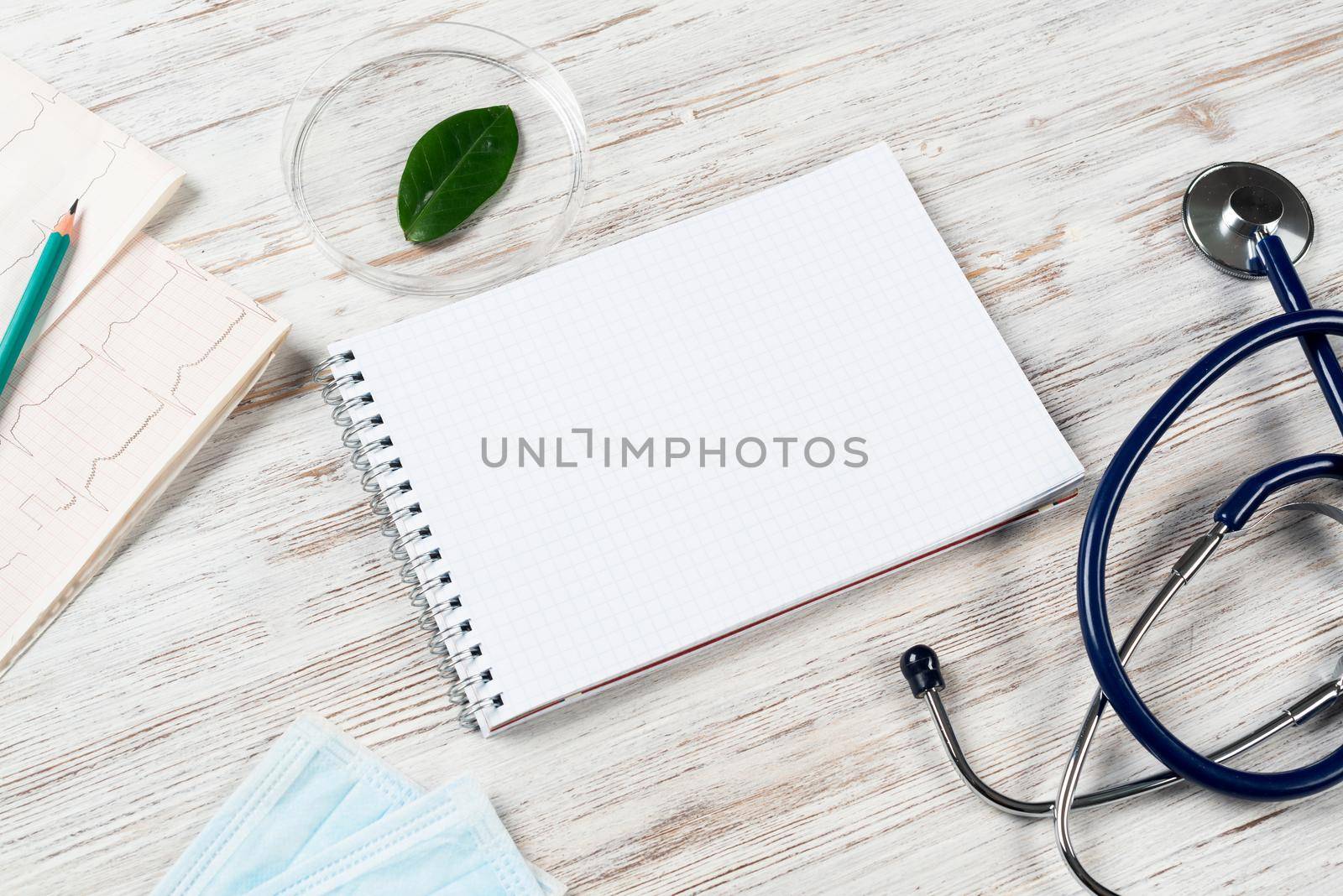 Patient examination and consultation concept with doctor workplace. Spiral notepad, disposable face mask, stethoscope and cardiogram on wooden surface. Professional healthcare in medical center.