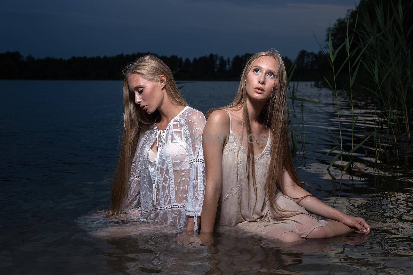 two attractive young twin sisters with long blond hair posing in light dresses in water of lake at summer night. stylish fashion photoshoot with flashlight. pretty models outdoors evening photosession