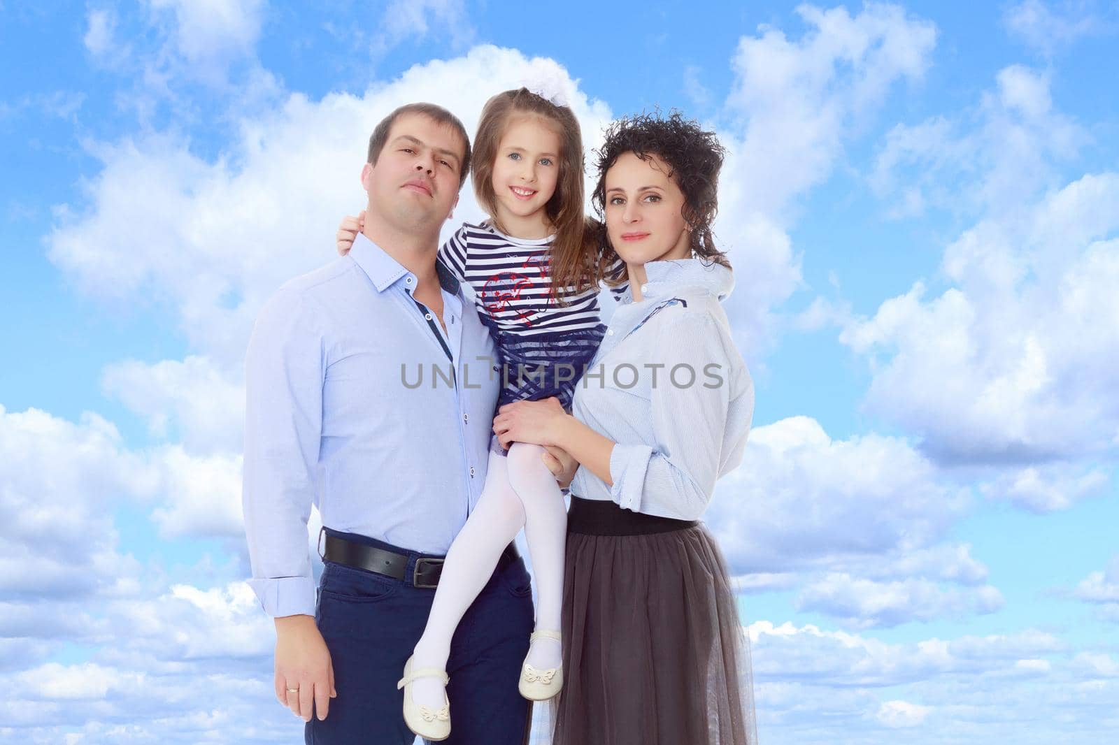 Happy young family, mom dad and little daughter.Parents keep the girl in her arms , and she hugs their neck.On the background of summer blue sky and fluffy clouds.
