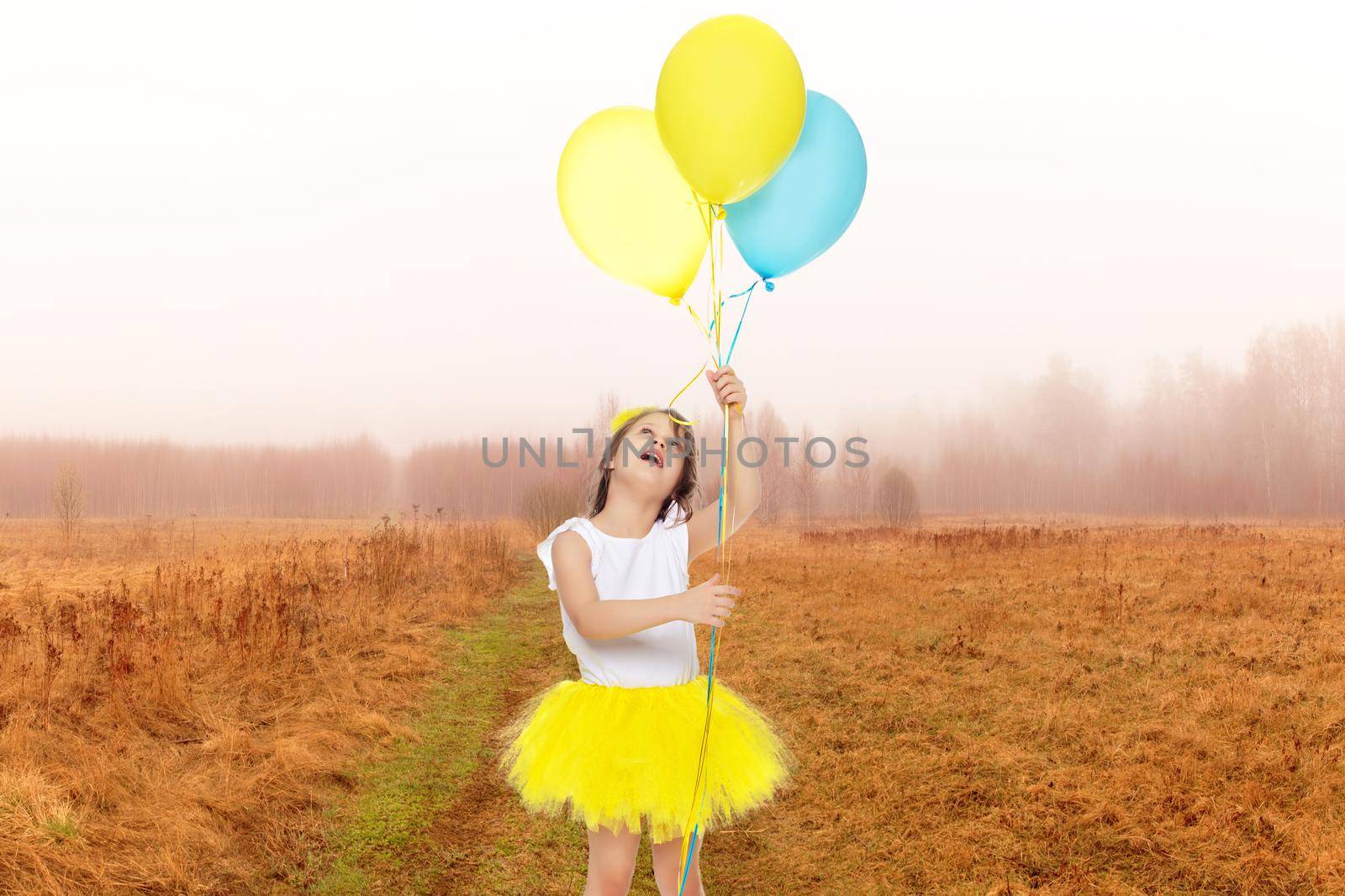 Pretty little blonde girl in a short bright yellow skirt and white blouse.Girl holds in hands balloons , She looks at them from the bottom up.Against the background of the summer landscape.