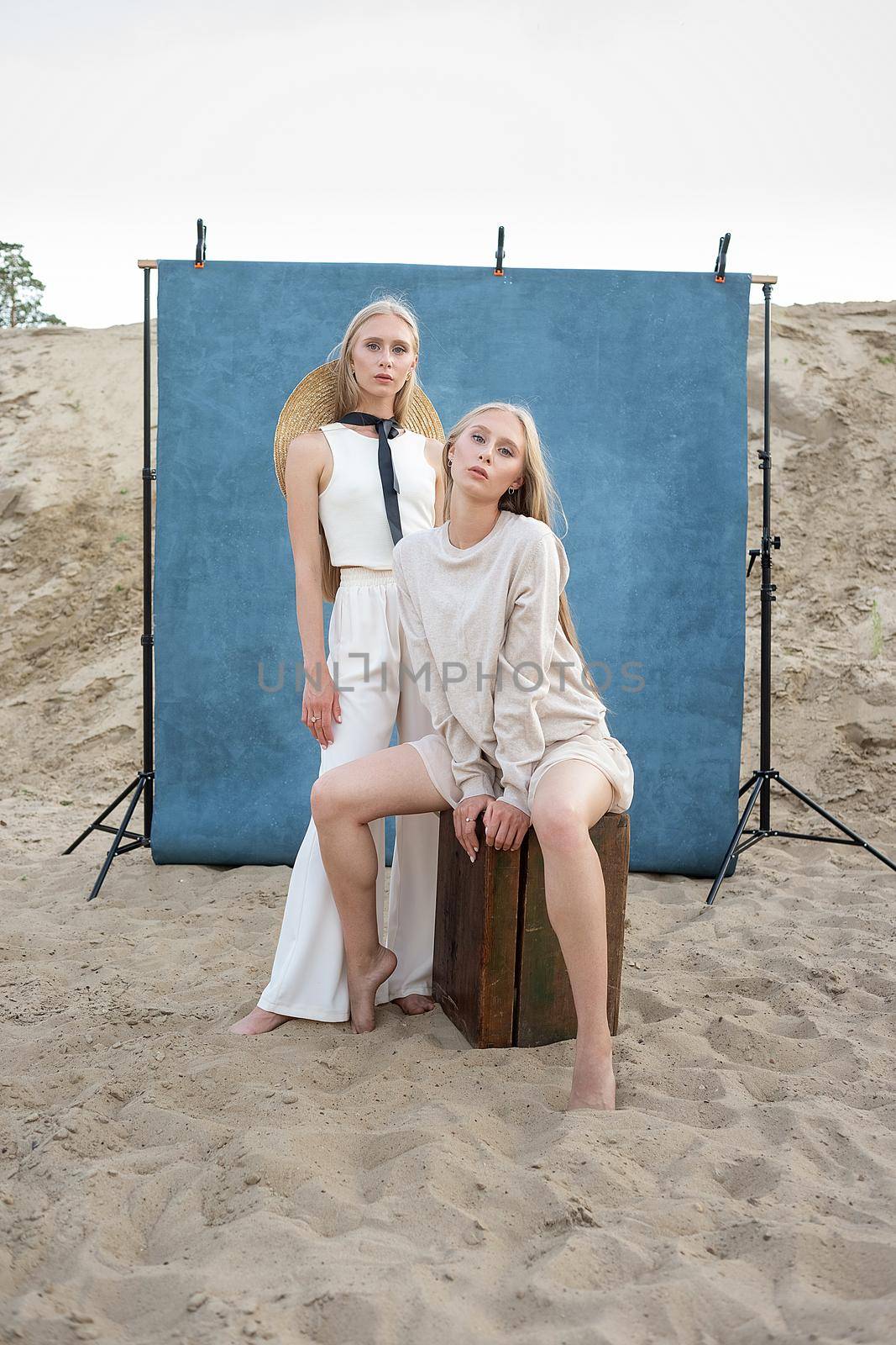 beauty portrait outdoors on sand in front of blue background, young pretty twins by artemzatsepilin