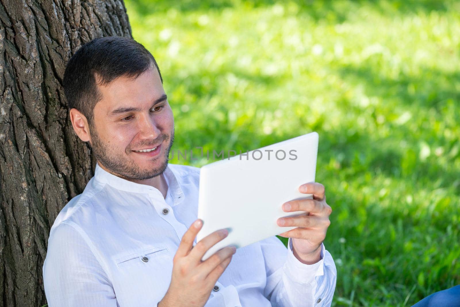 Man using tablet computer under tree in park on sunny day. Handsome guy looking at tablet screen. Happy smiling man sitting on green grass leaning against tree. Digital technology and communication
