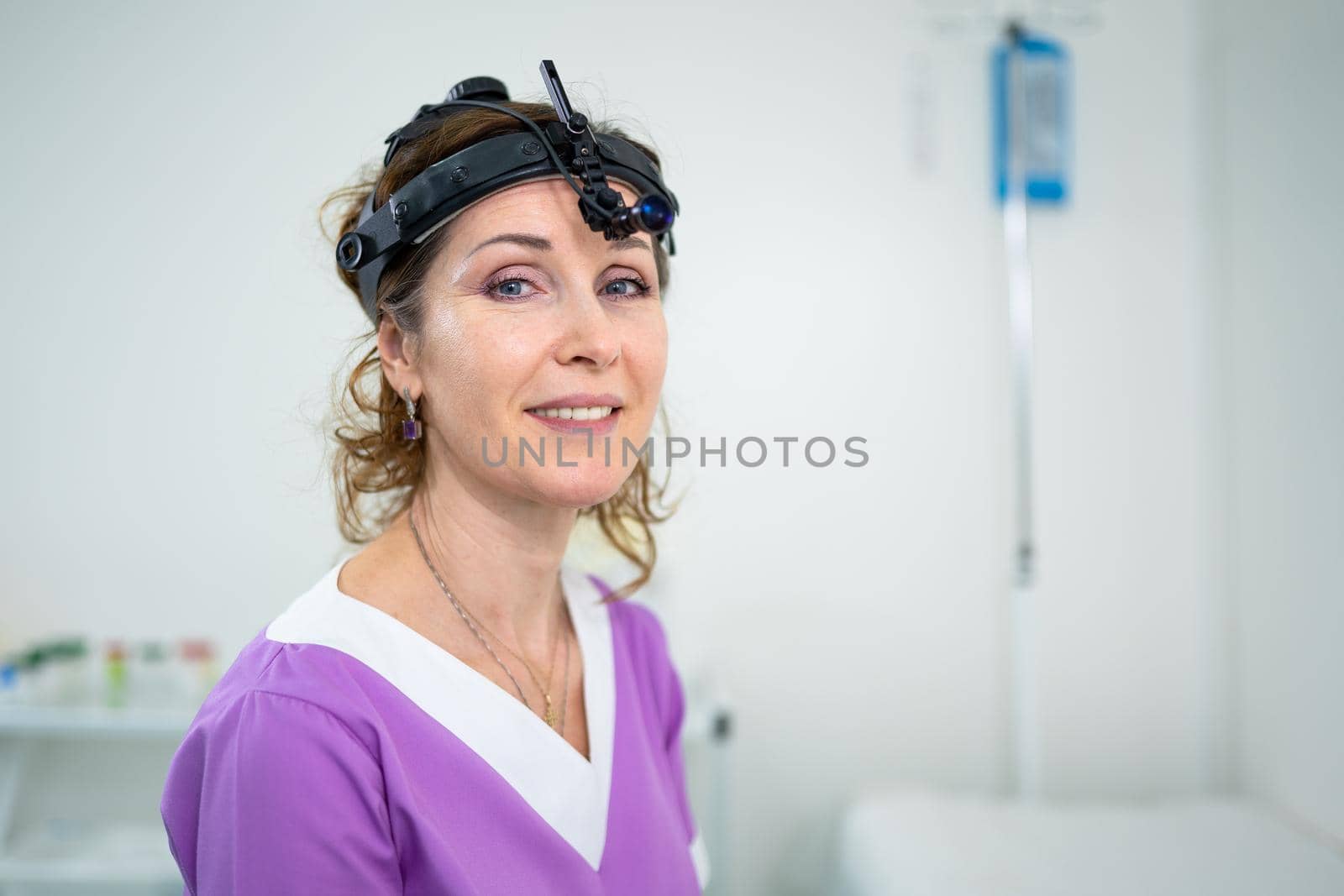 Medical worker of ENT clinic middle aged Caucasian female in purple medical uniform posing looking at the camera in the examination room. Professional medical specialist otolaryngologist in hospital by Tomashevska