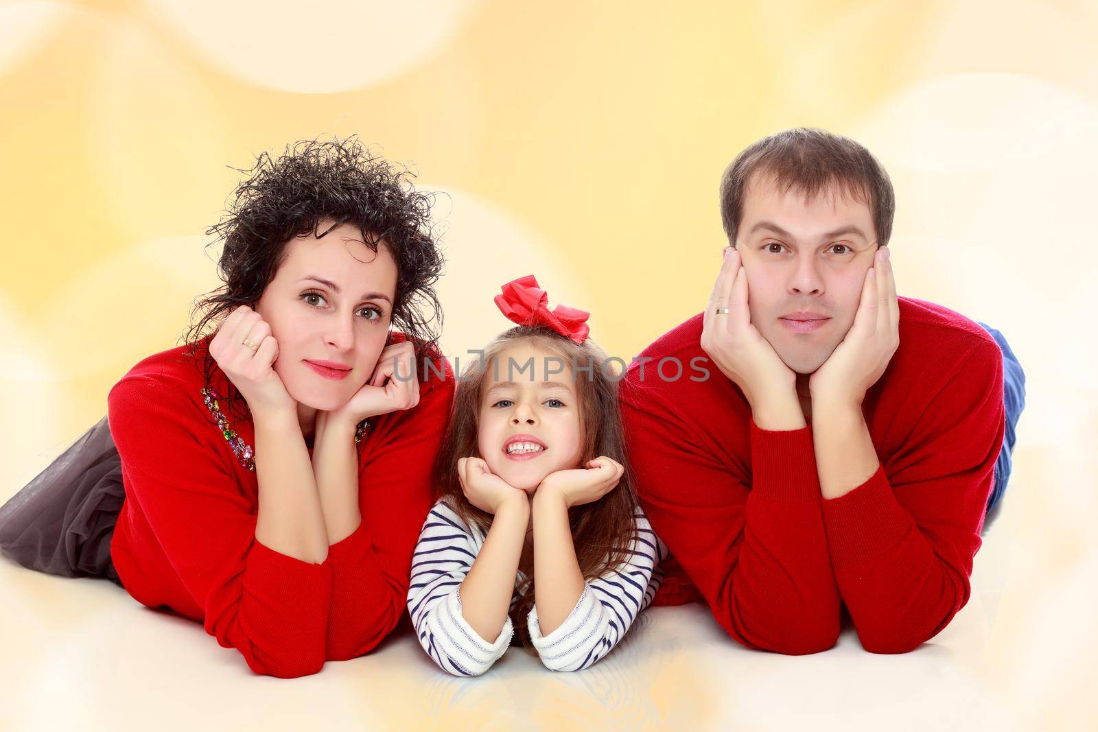 Happy young family dad mom and a little girl in bright red outfits . Family lying on the floor leaning on his hands.Brown festive, Christmas background with white snowflakes, circles.