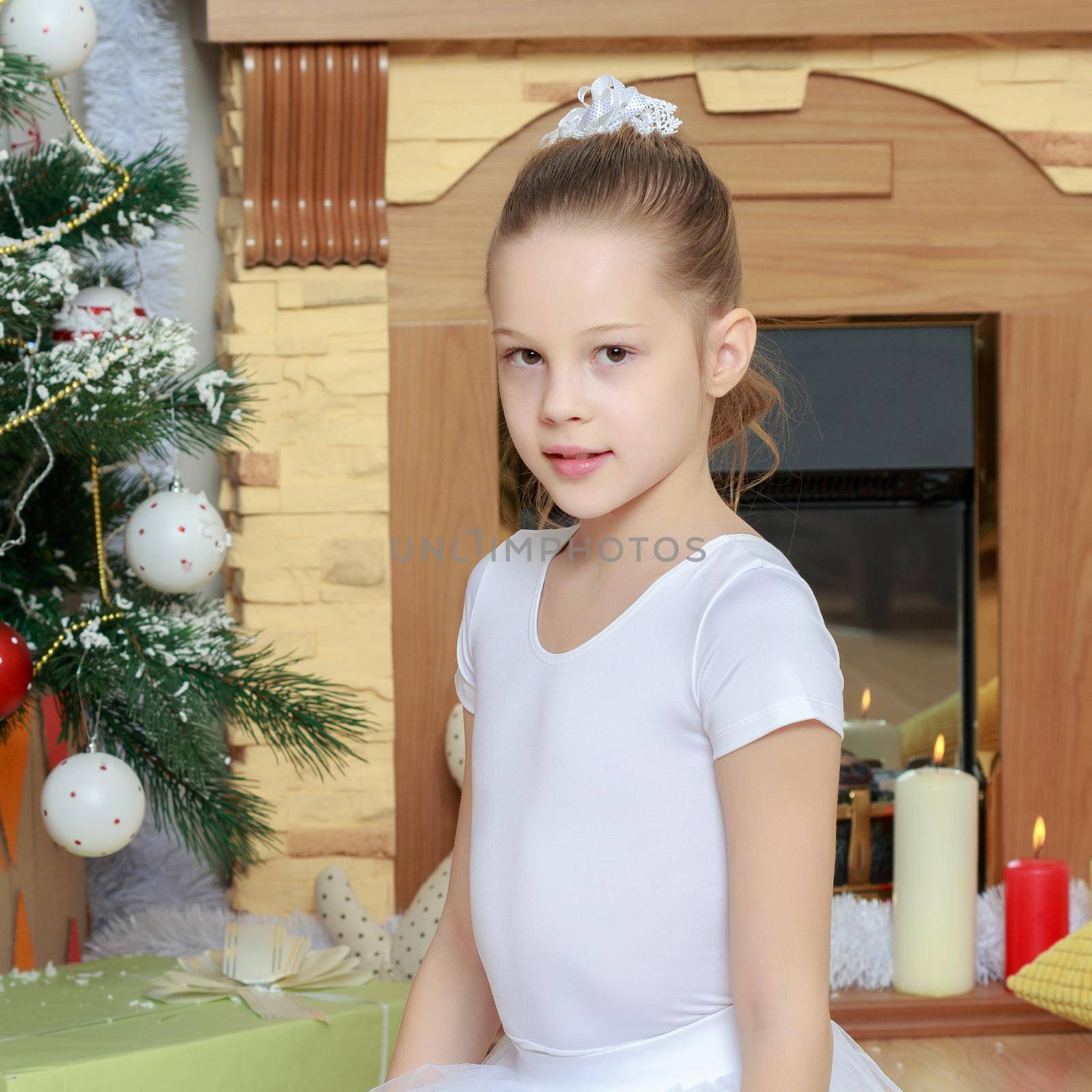 Cute little girl in smart white dress Christmas around the fireplace which burn holiday candles.