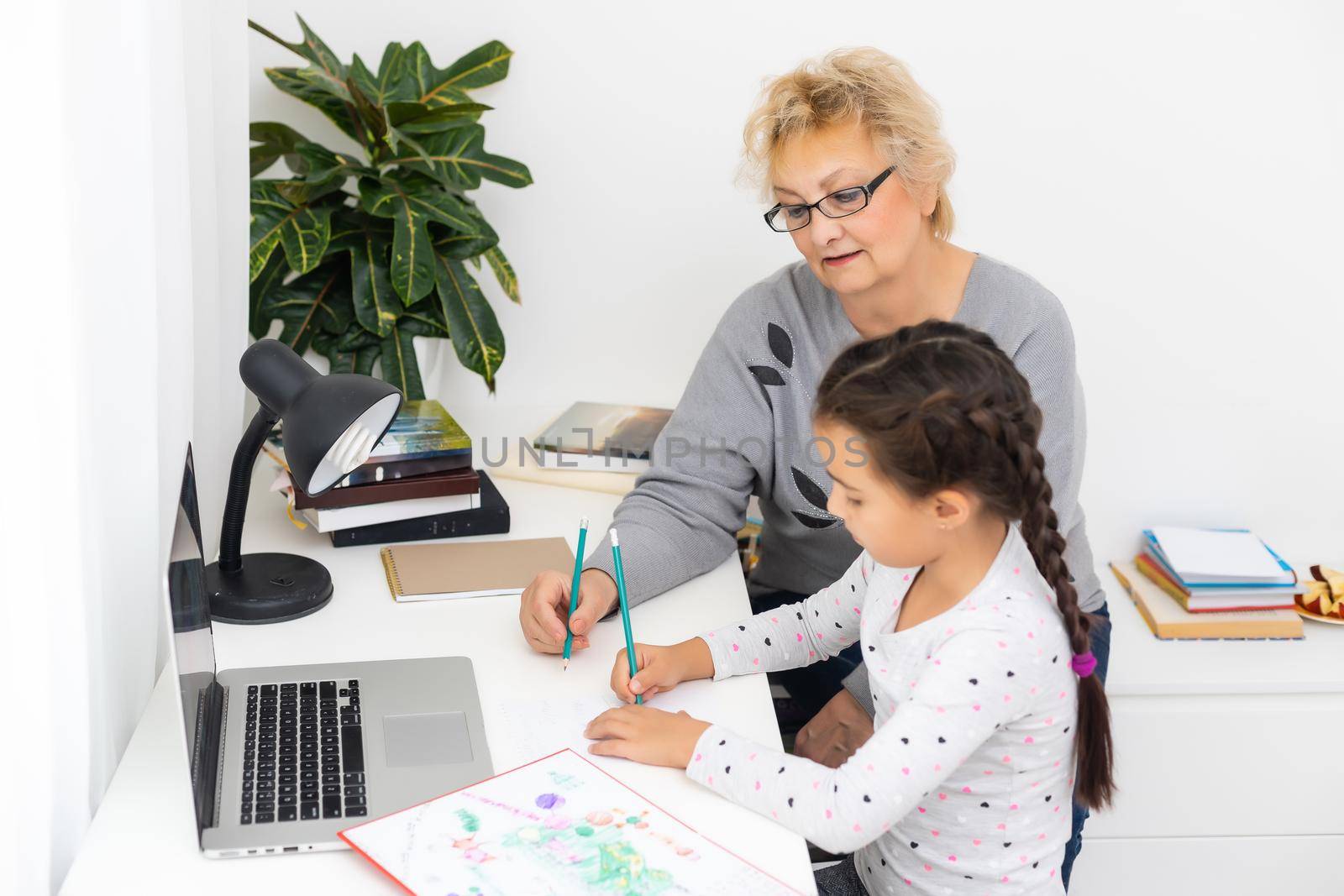 Mature grandmother helping child with homework at home. Satisfied old grandma helping her granddaughter studying in living room. Little girl writing on notebook with senior teacher sitting next to her by Andelov13