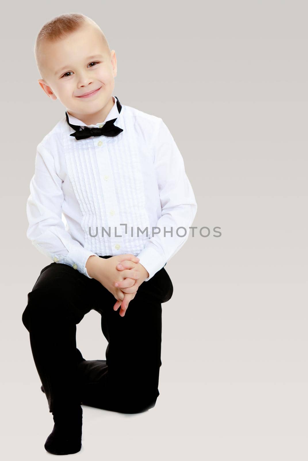 Elegant little boy in black pants ,white shirt and black tie. Standing on one knee.On a gray background.
