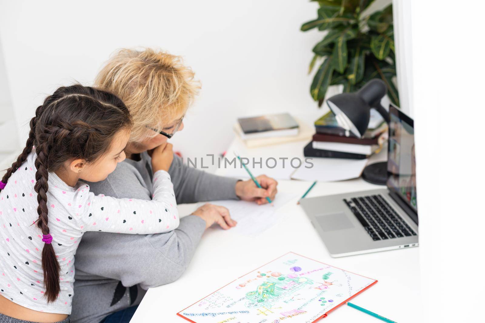 Mature grandmother helping child with homework at home. Satisfied old grandma helping her granddaughter studying in living room. Little girl writing on notebook with senior teacher sitting next to her