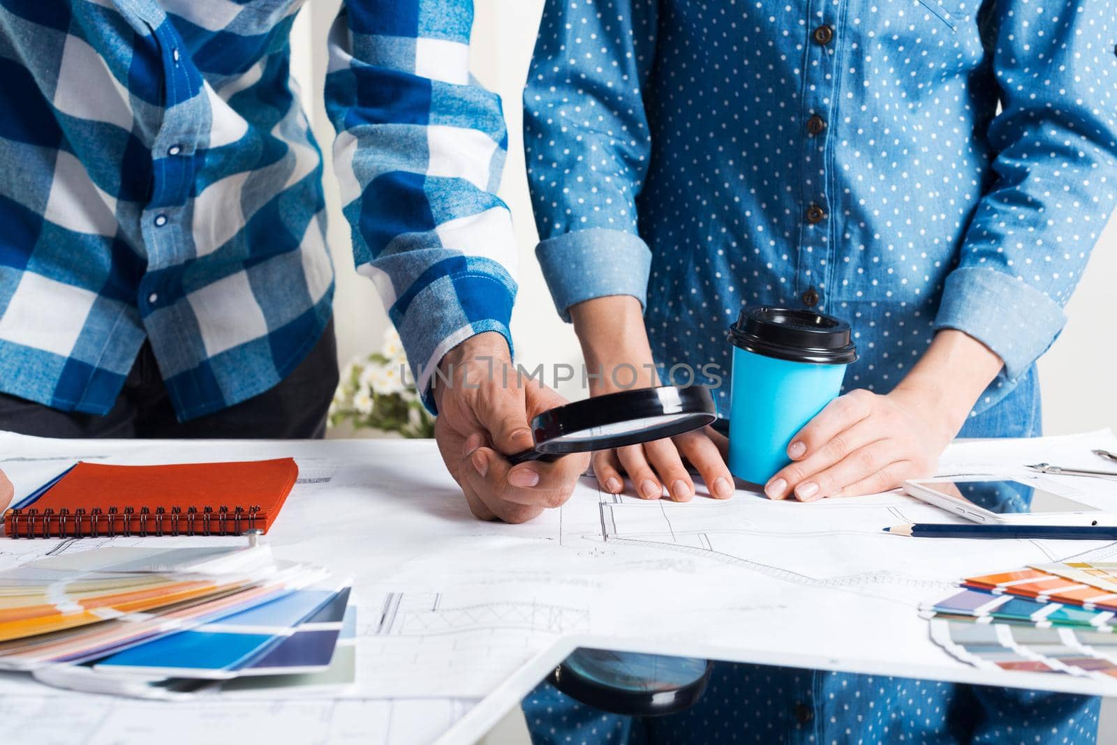 Man carefully studying technical drawing with magnifying glass. Designers working with color swatches and construction blueprint at workplace. Thorough analysis and inspection of design project.