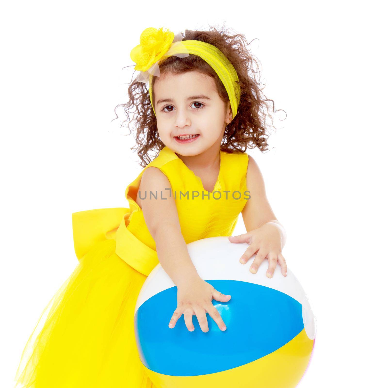 Funny curly baby girl in a bright yellow dress and bow on her head playing with a ball.Isolated on white background.
