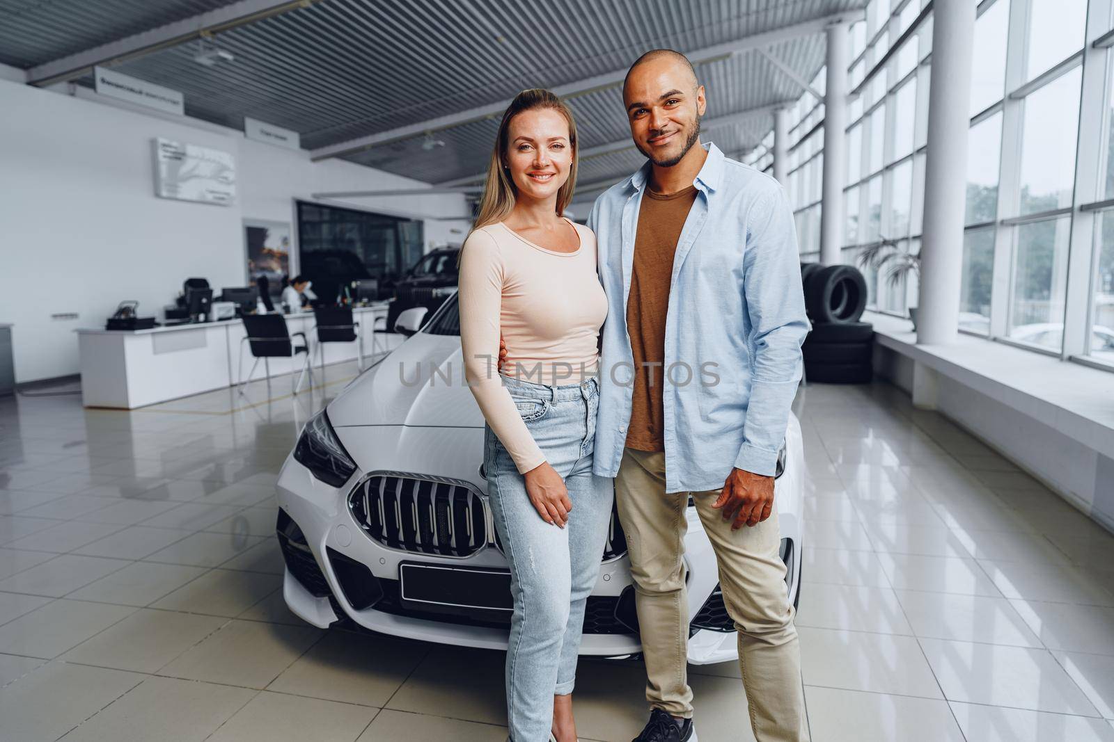 Happy couple of caucasian woman and african american man standing near their new luxury car inside car salon