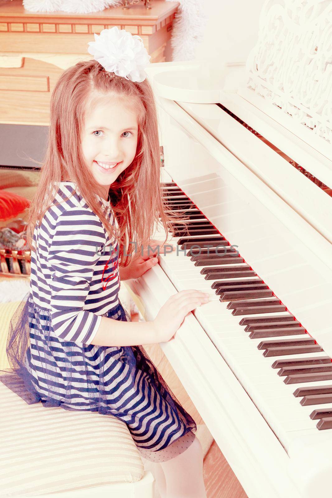 Little girl in blue striped dress and a white bow on her head.Girl smiling sitting behind the keys of a large white piano.Creative toning of a photograph.