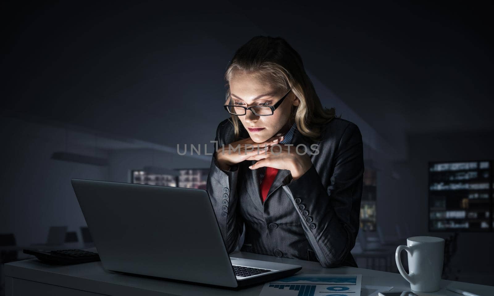 Portrait of concentrated businesswoman in glasses sitting at office desk and working at laptop. Financial consultant in business suit and tie busy at work in evening. Working in office late at night.
